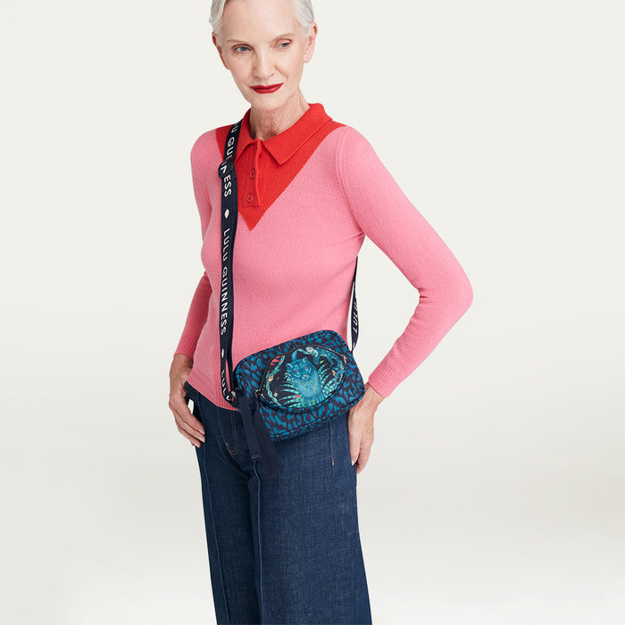 Woman wearing a pink sweater with a red collar and blue jeans, holding a patterned Lulu Guinness x Emma Shipley crossbody bag.