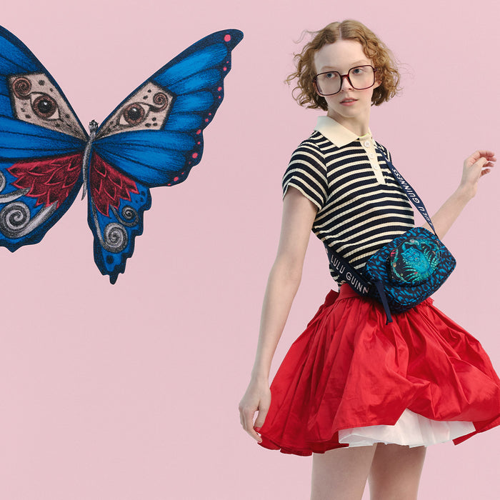 A girl with curly red hair and glasses is shown wearing a black and white striped t-shirt, a bright red skirt, red-and-white striped socks, and red shoes. She is wearing a blue patterned crossbody bag.