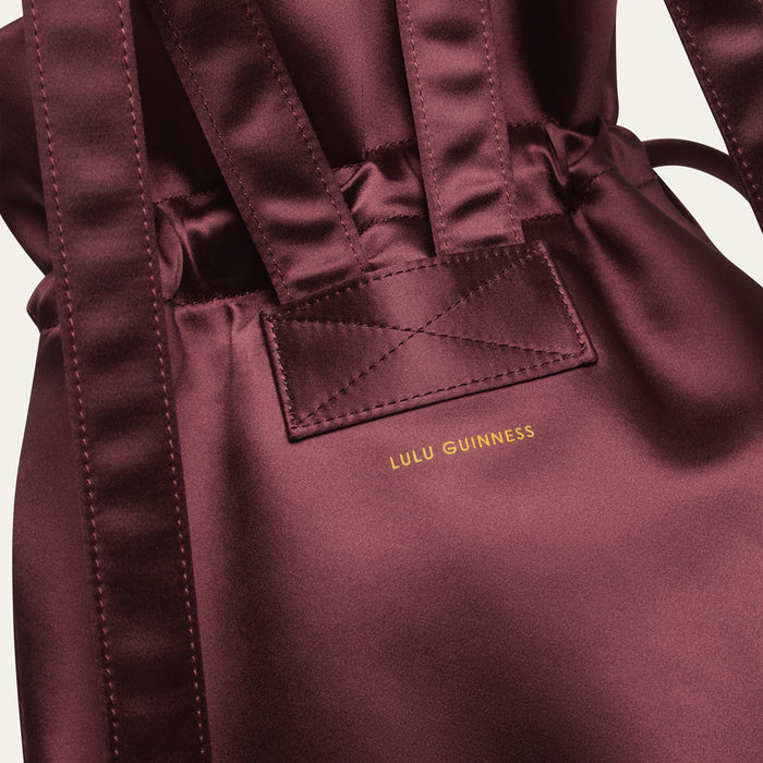 Maroon satin backpack with 'Lulu Guinness' logo on a light gray background