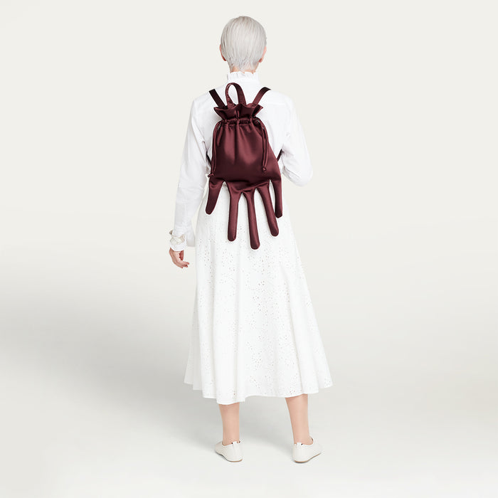 Person wearing a white dress and carrying a maroon satin hand-shaped backpack on a plain background