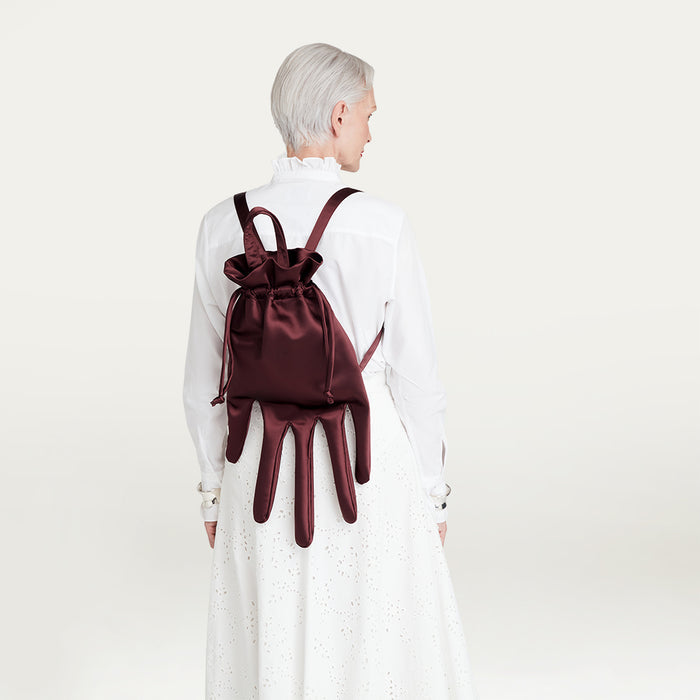 Person wearing a white outfit with a burgundy satin hand-shaped backpack on a plain background
