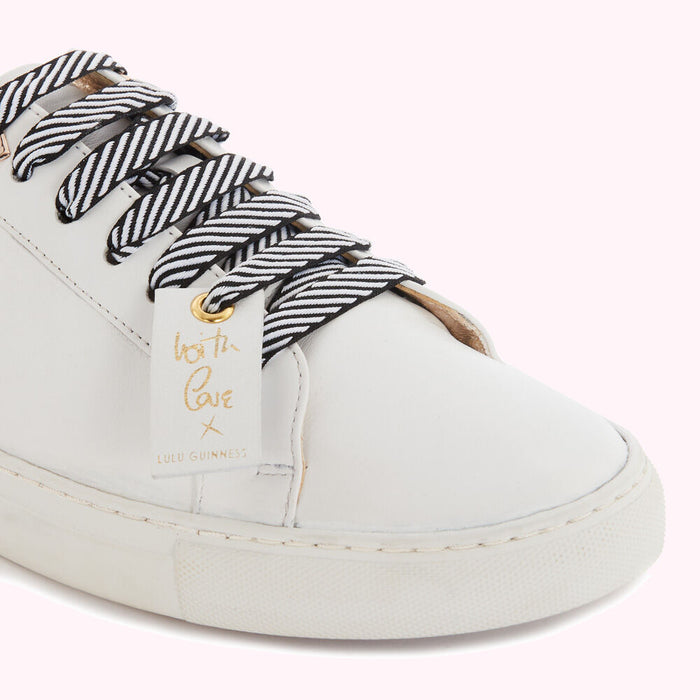 Close-up of white leather Lulu Guinness sneakers with black and white striped laces and a gold "With Love" tag.