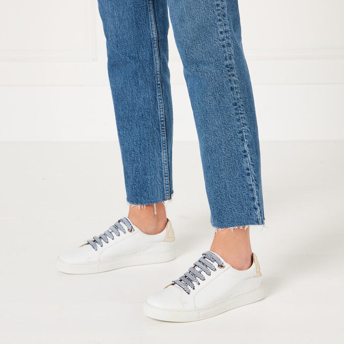Person wearing white sneakers with navy and cream striped laces and blue jeans against a white background.