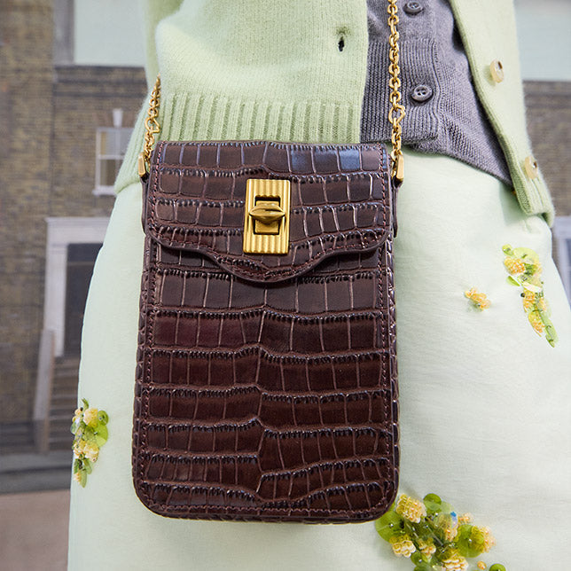A dark brown croc-effect embossed leather crossbody bag with a gold clasp and chain strap, worn over a light green and gray cardigan.