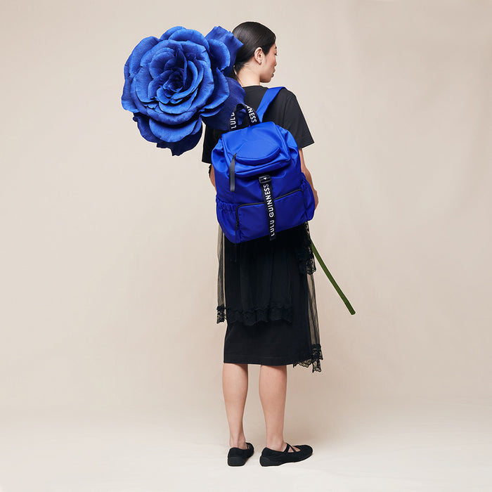 Person wearing a blue backpack with a large blue flower held above them against a beige background