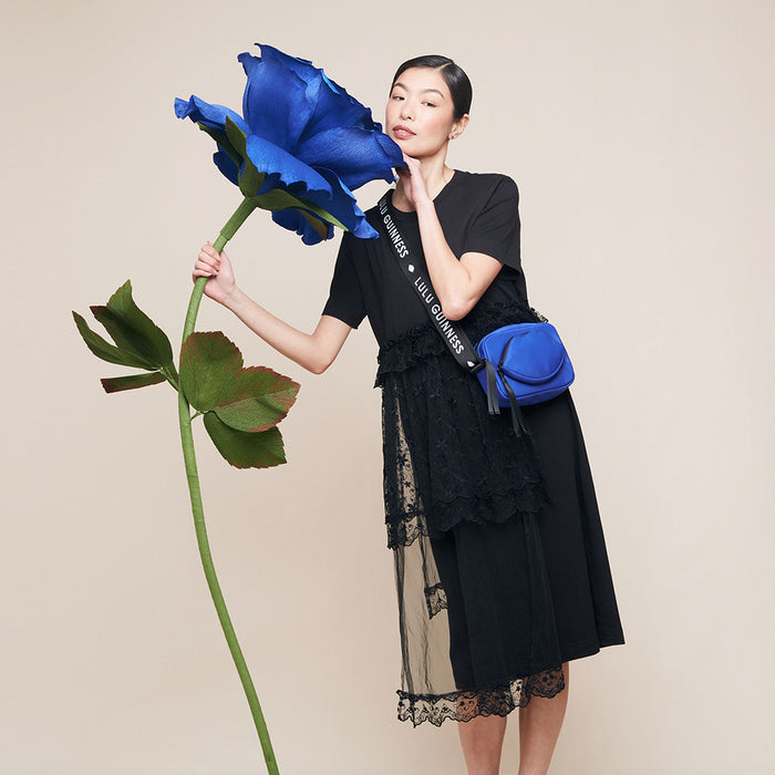 Woman holding a large blue flower against a beige background and wearing a blue nylon crossbody bag with a 'Lulu Guinness' branded strap