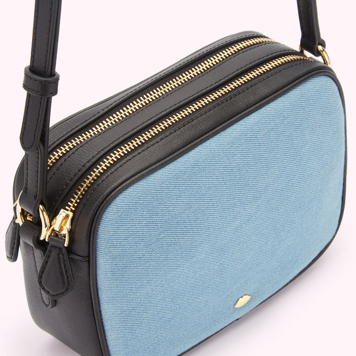 Close-up of a light blue denim crossbody bag with black leather trim and gold zippers.