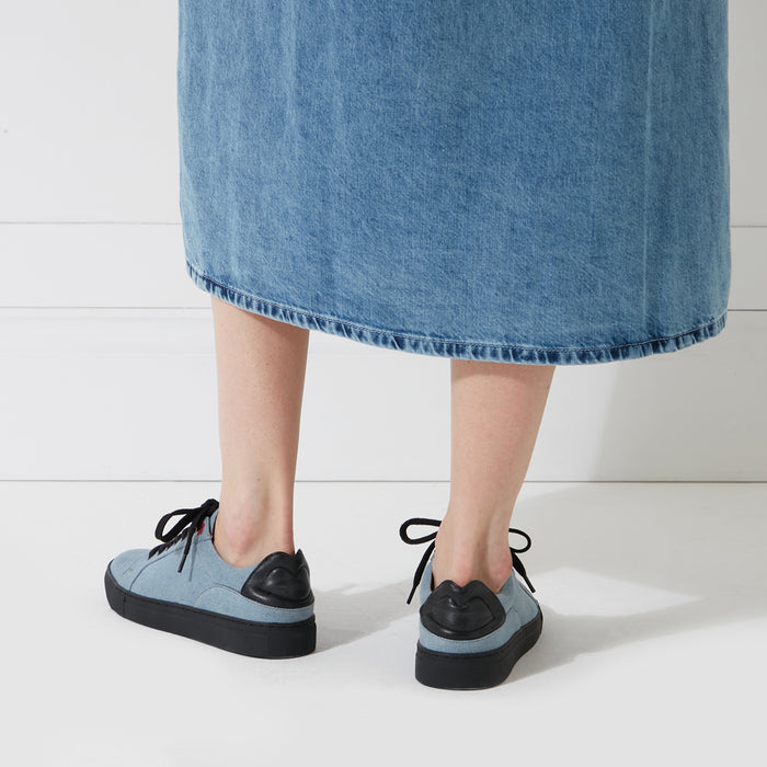 Person wearing light blue denim midi skirt and light blue canvas sneakers with black lip detail on the back. The background is white.