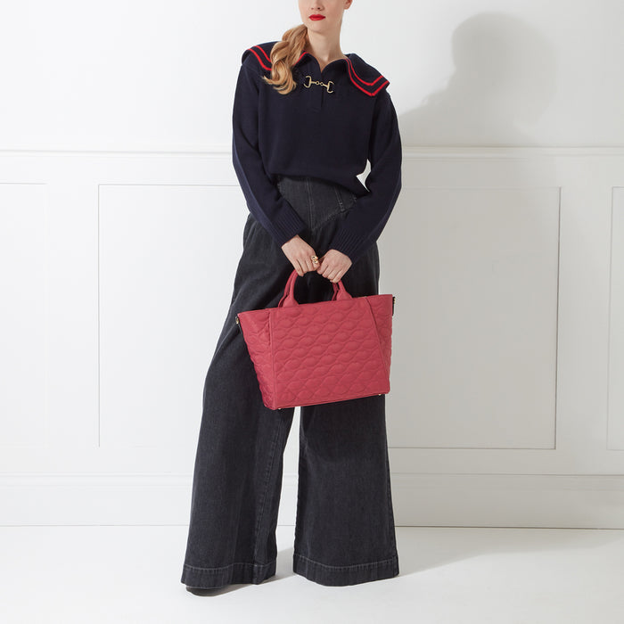 A woman in dark-wash wide-leg jeans and a navy sweater with a sailor-style collar holds a quilted pink tote bag against a white background.