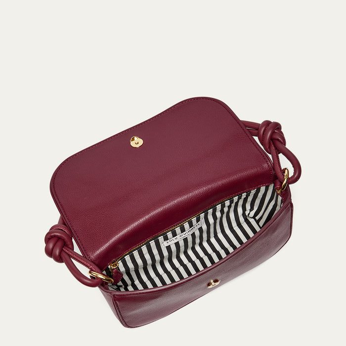 Inside of a burgundy leather shoulder bag revealing a black and white stiped lining, inside pocket and gold accents on a light grey background