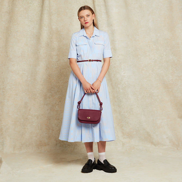 Woman in a light blue dress holding a burgundy leather handbag against a beige background