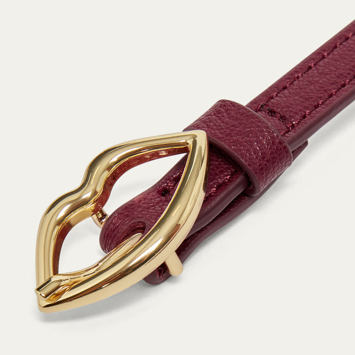 Close up image of a burgundy leather belt with a gold lip shaped buckle on a light grey background