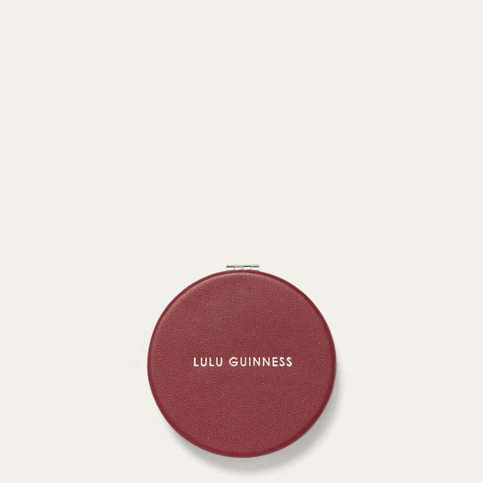 Round burgundy red compact mirror with 'Lulu Guinness' branding on a light beige background
