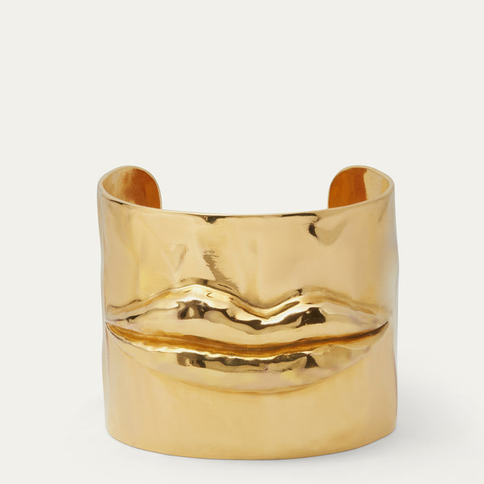 A polished gold-tone cuff bracelet shaped like a pair of lips, photographed on a clean off-white background.