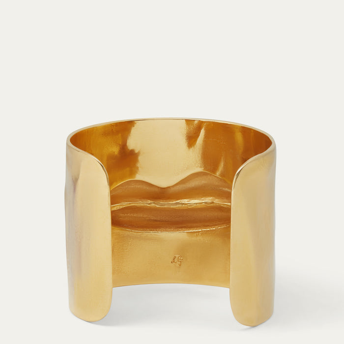 A wide, open cuff bracelet in polished yellow gold with a smooth, slightly wavy surface, photographed on a clean white background.