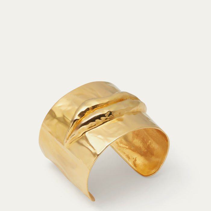 A wide, hammered gold cuff bracelet with a textured, organic lip design, photographed on a clean off-white background.
