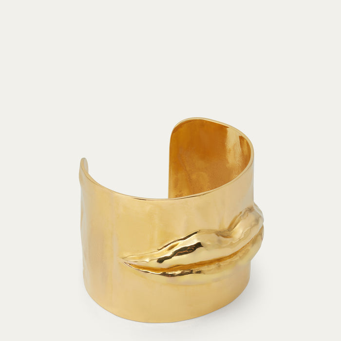 A polished gold-tone cuff bracelet with a sculptural, lip detail on the front, photographed against a clean off-white background.