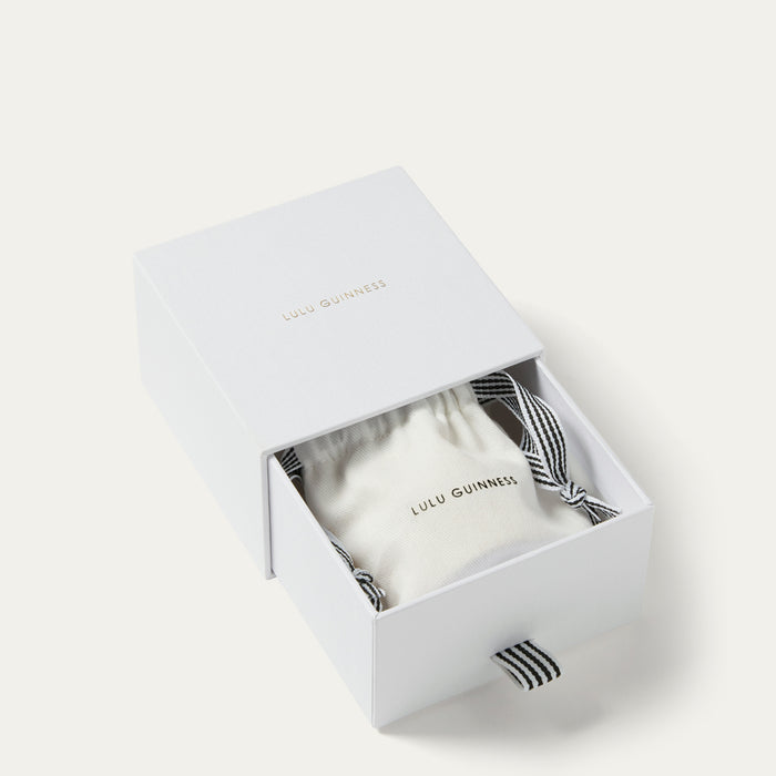 An open white Lulu Guinness gift box with its lid propped open, revealing a white drawstring pouch inside. The box lid has gold foil branding, and the drawstring bag has a black-and-white striped ribbon.