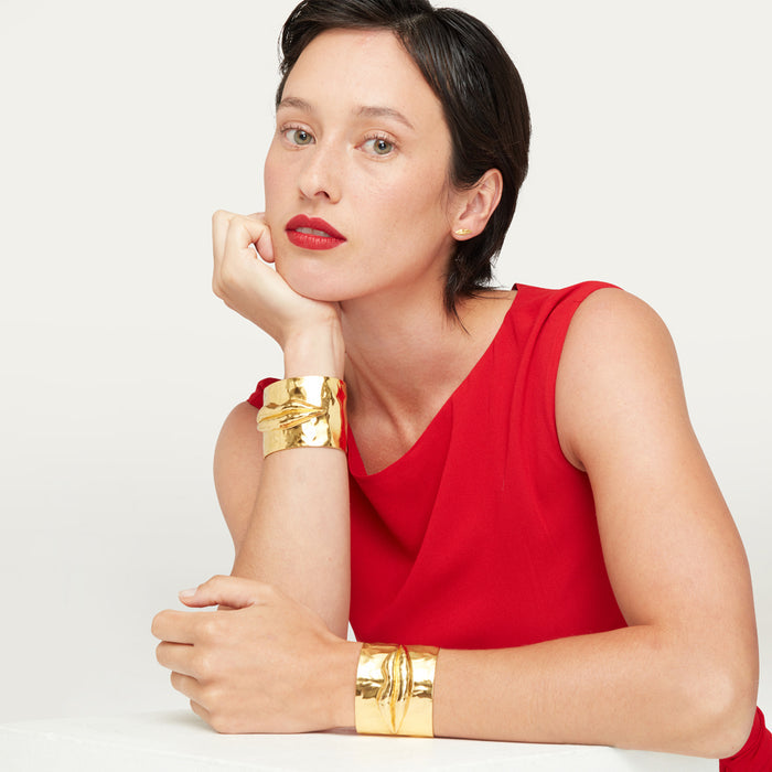A women with dark short hair wearing a red dress resting on a plinth wearing two gold cuff bracelets.