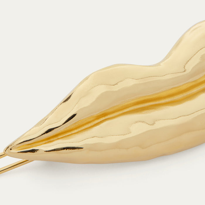 A close-up of a gold-toned, lip-shaped hair clip on a clean off-white background.
