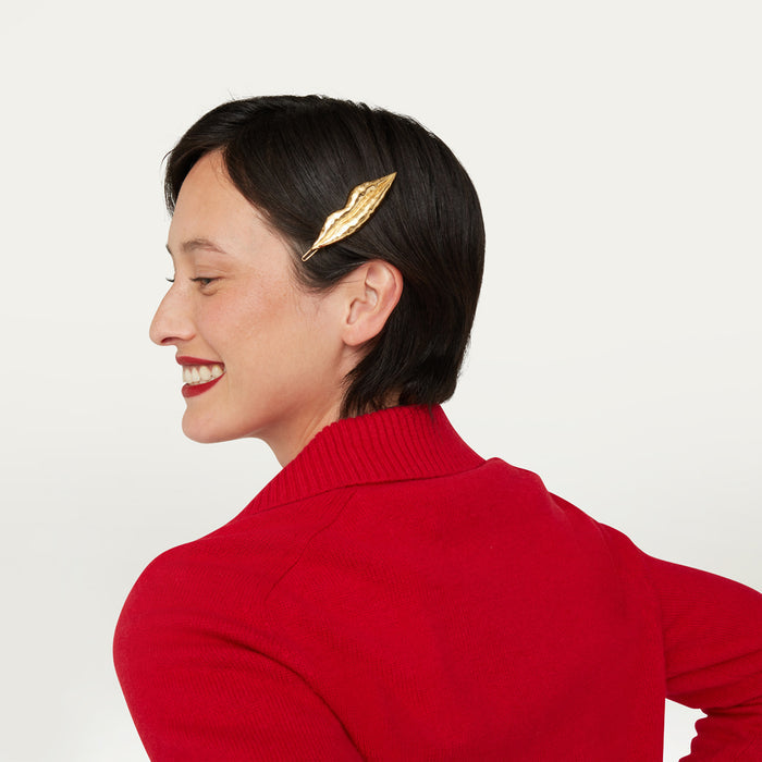 A woman with short dark hair is shown in profile wearing a gold lip-shaped hair clip on the side of her head, paired with a bright red ribbed turtleneck sweater against a clean off-white background.