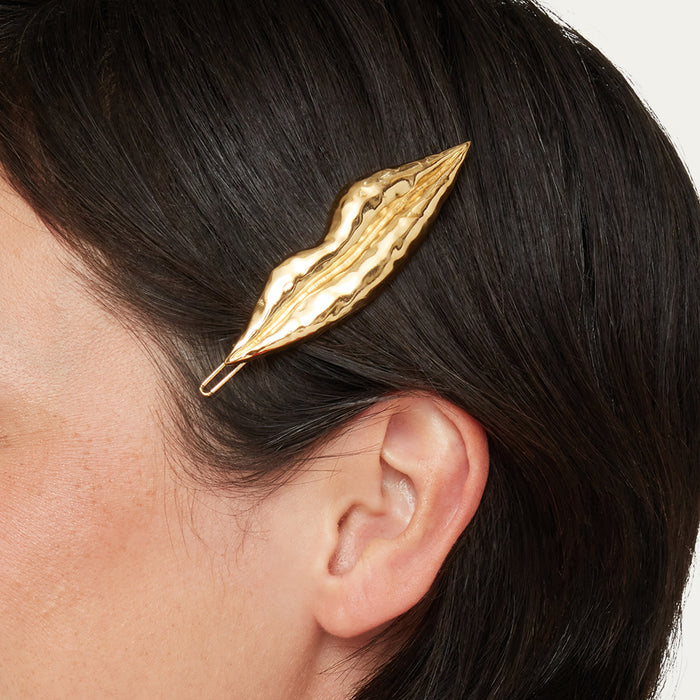 A gold lip-shaped hair claw clip with a textured, wavy surface, shown worn on a person's dark hair.
