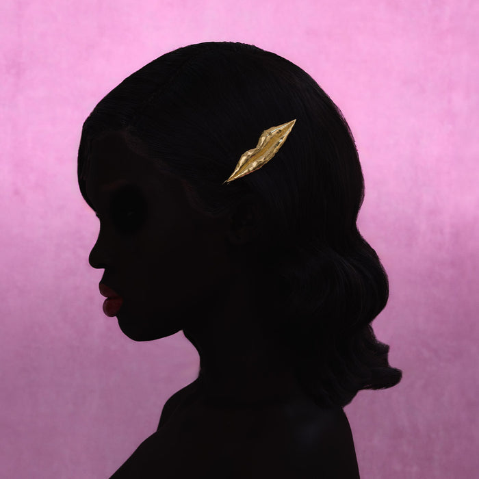 A single gold lip-shaped hair clip, shown on a womans profile against a textured pink background.