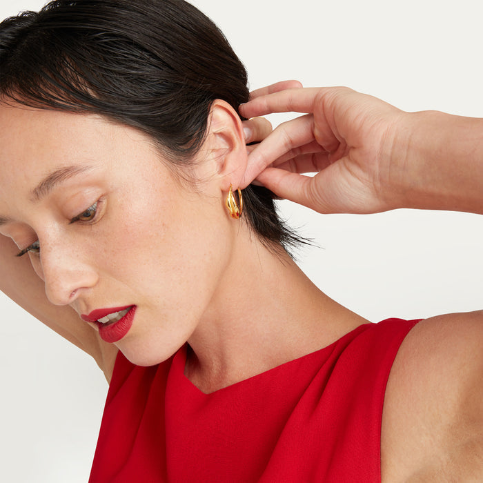 A close-up lifestyle shot of a woman wearing a small gold hoop earring, with her hand adjusting the earring on her ear. She is wearing a bright red sleeveless top against a clean, light off-white background.