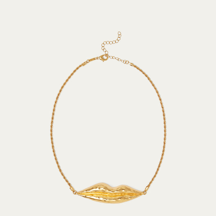A gold necklace featuring a textured, sculptural lip-shaped pendant suspended from a delicate gold chain, set against a solid off-white background.