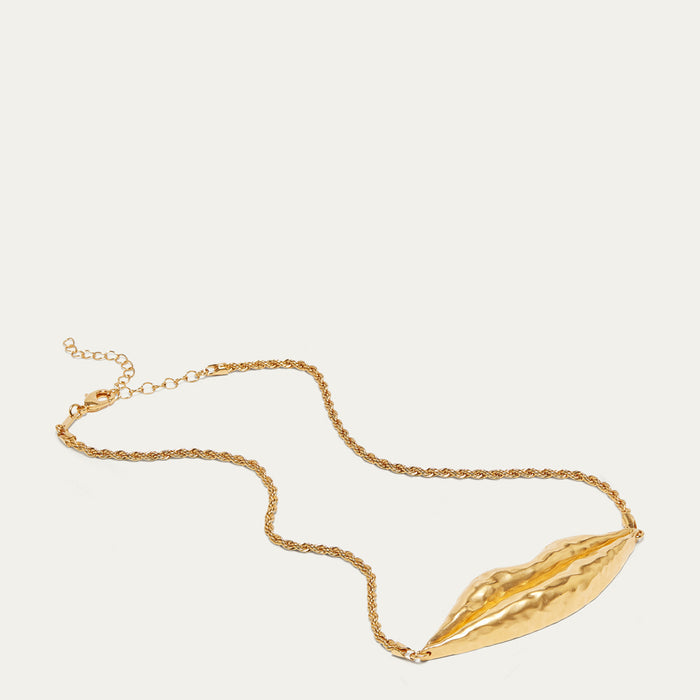 A gold necklace featuring a textured, sculptural lip-shaped pendant on a delicate cable chain, displayed against a solid off-white background.