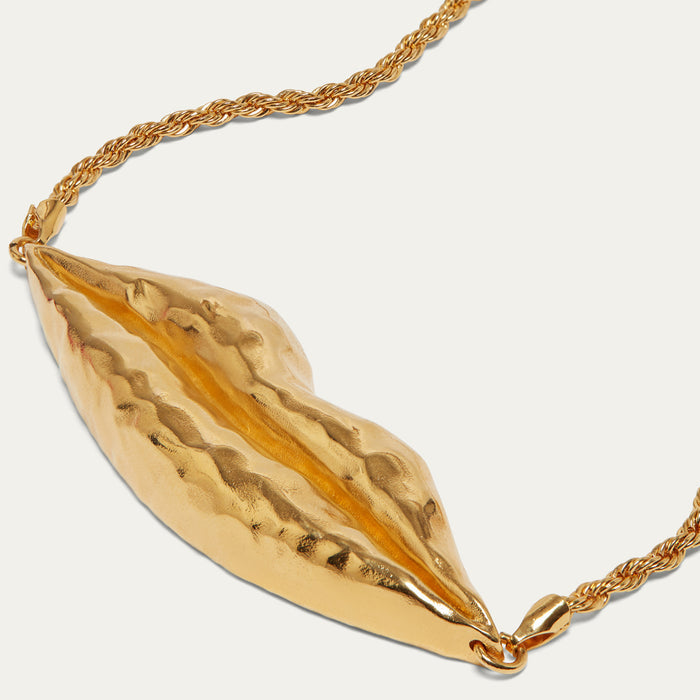 A gold-toned necklace featuring a textured, hammered metallic pendant shaped like stylized lips, suspended from a twisted chain on a light off-white background.
