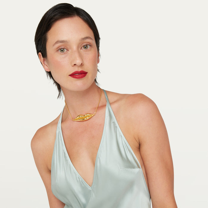 A woman with short dark hair and red lipstick wears a gold necklace with a sculptural lip-shape pendant, paired with a pale mint green satin halter top, against a plain off-white background.