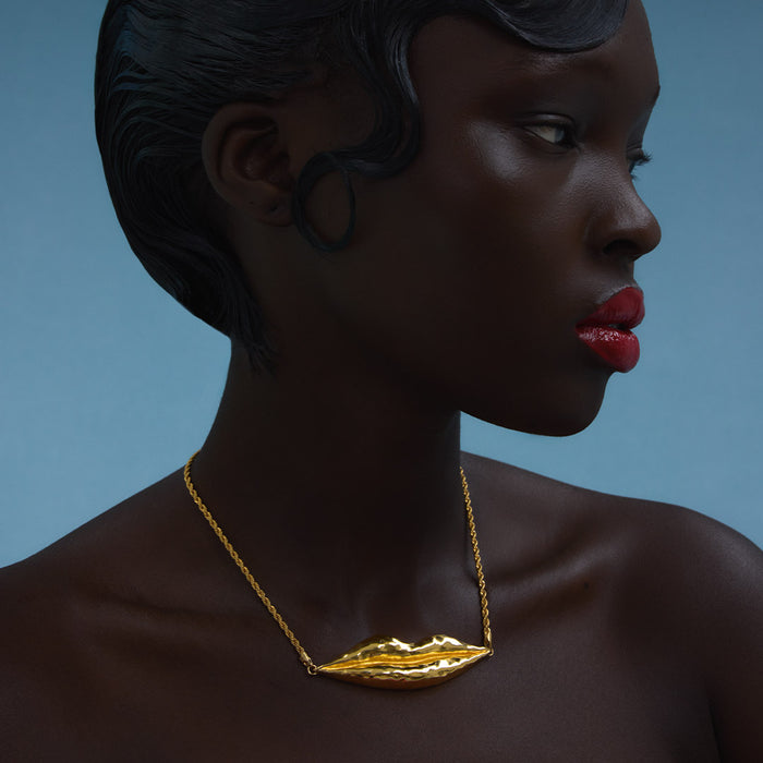A close-up side profile of a model wearing a textured gold necklace shaped like lips, set against a solid blue background.