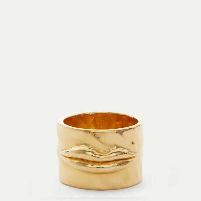 A wide gold ring featuring a sculpted, textured lips motif on the front, photographed against a clean, light off-white background.