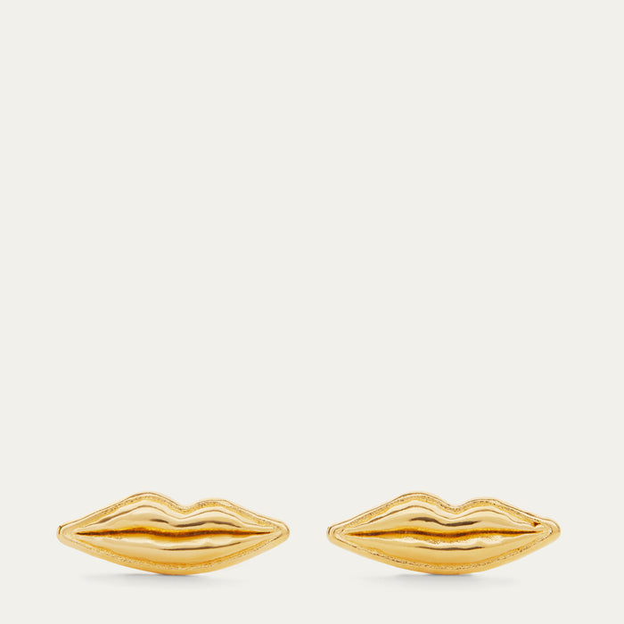 A pair of gold lips-shaped stud earrings on a light cream background.