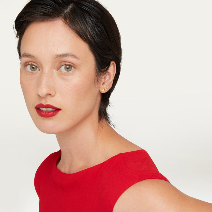 A close-up of a woman with short dark hair wearing a bright red top and a small gold lip-shaped stud earring, photographed against a clean light gray background.