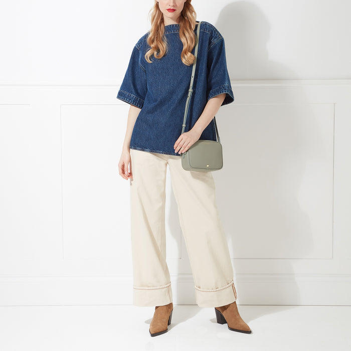 A woman in a denim shirt and cream pants models a sage green crossbody bag against a white background.
