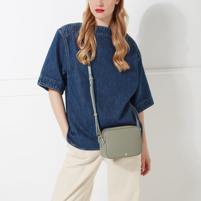 A woman in a denim shirt and beige pants wears a pale green crossbody bag.