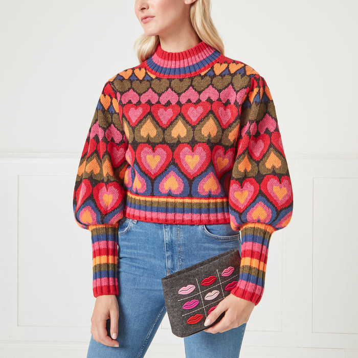 Woman in colourful heart-patterned sweater holding grey clutch with lips design.