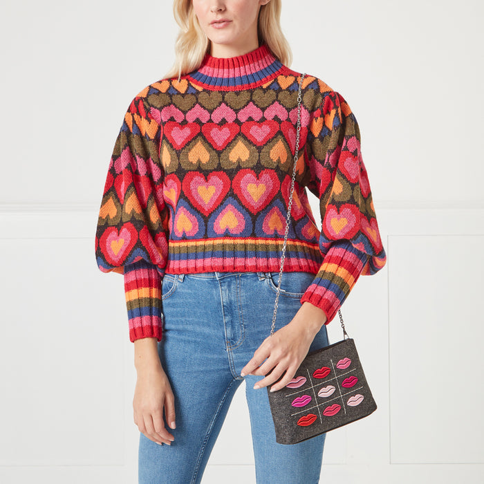 A woman in a multi-coloured heart patterned sweater and blue jeans holds a grey crossbody bag adorned with lips.