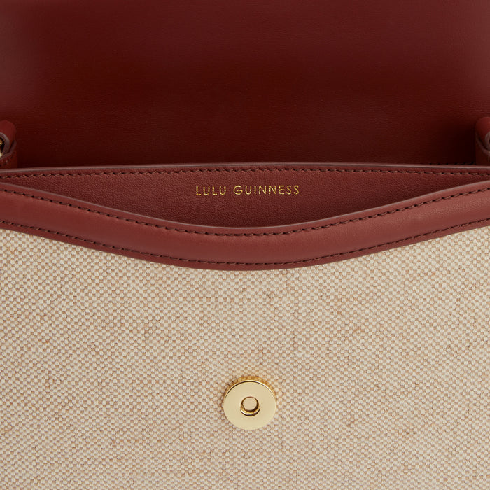 Close-up of the inside of a Lulu Guinness bag, showing the beige canvas body, burgundy leather trim, and gold-toned press stud. The Lulu Guinness logo is visible inside.