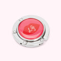 Silver handbag hook with a red circular design featuring a lip print and the letters L.G.J.