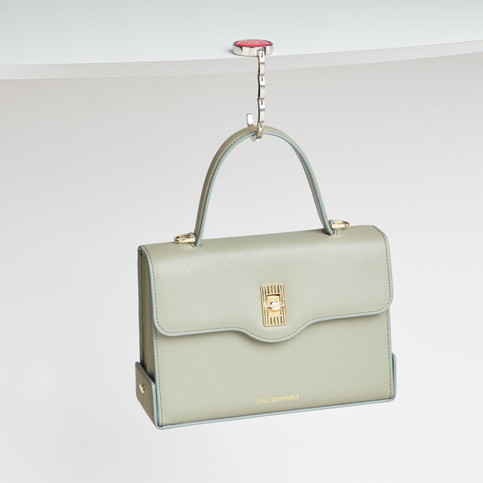 A pale green Lulu Guinness handbag hangs from a silver hook attached to a white surface.