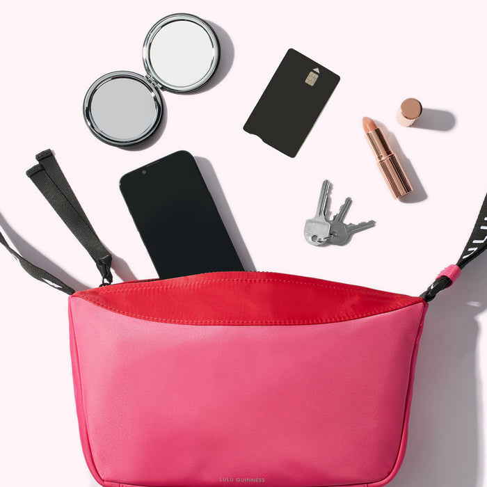 A bright pink and red nylon crossbody bag with a smartphone, keys, compact mirror, card and lipstick spilling out.