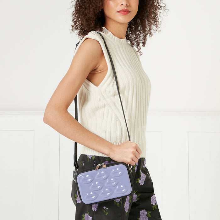 Woman wearing a cream-colored sleeveless sweater and black floral shorts holds a lavender crossbody bag with a black strap and lips embossed on the front. The background is white.