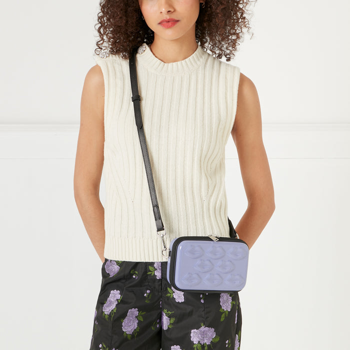 A woman with curly brown hair is modelling a lavender crossbody bag with a black strap and embossed lips pattern, worn over a cream-colored ribbed sleeveless top and black floral shorts. The background is white.