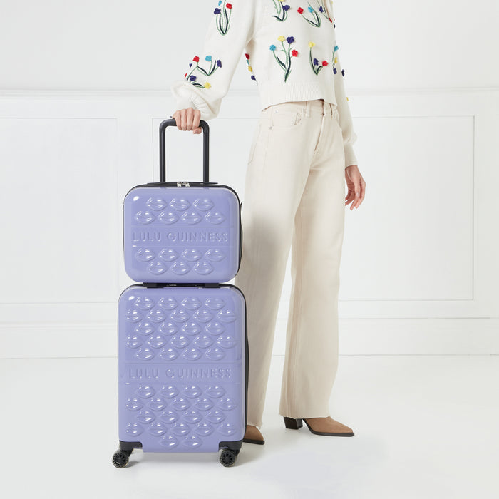 A woman in beige pants and a white embroidered sweater stands beside a lilac Lulu Guinness two-piece suitcase set with a kiss print.