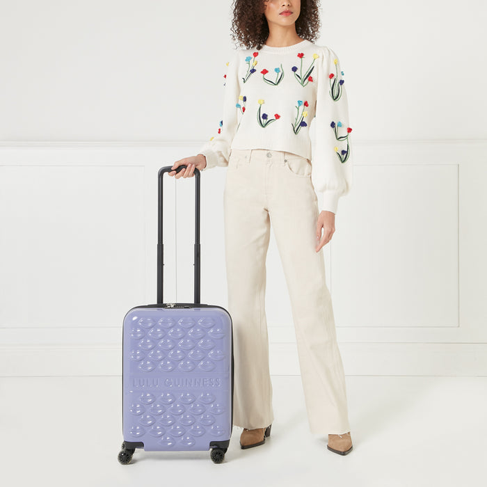 A woman in a cream floral sweater and beige pants stands holding a purple Lulu Guinness suitcase with a lip pattern. The background is white.