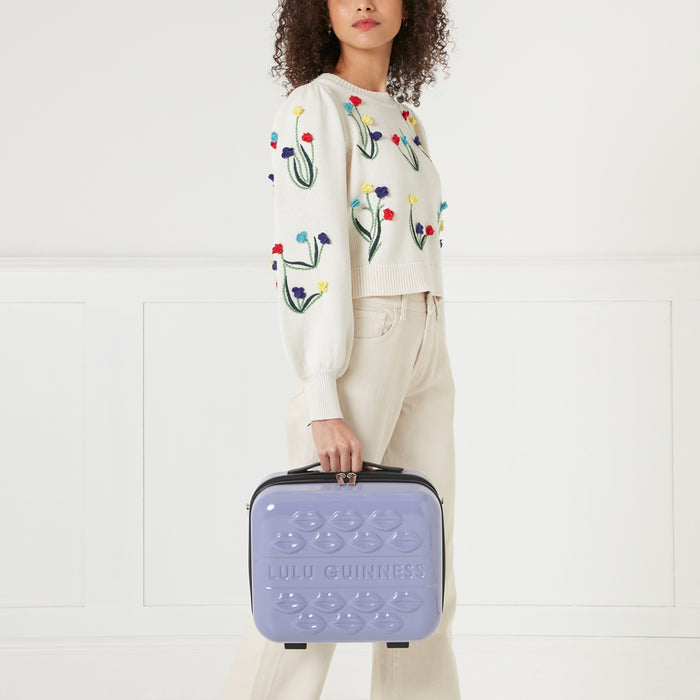 A woman in a floral embroidered cream sweater and beige pants holds a lavender Lulu Guinness makeup case adorned with embossed lips.