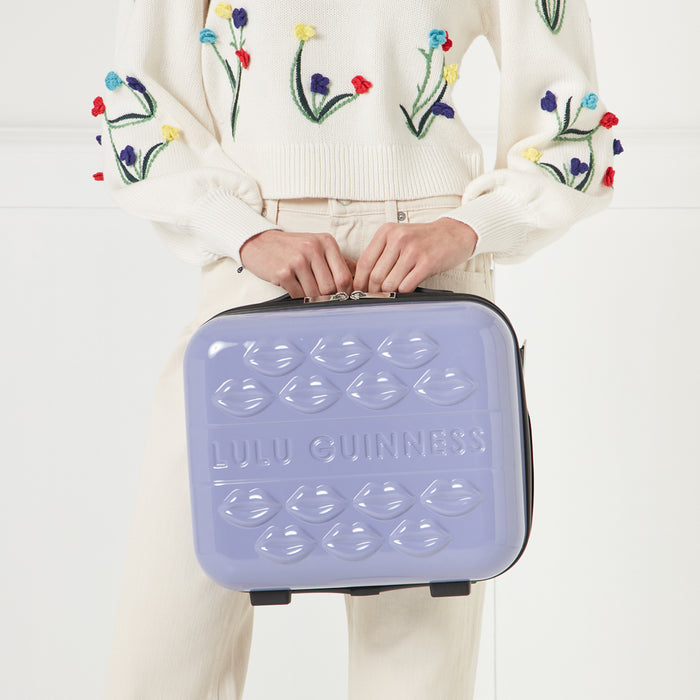 A woman in a cream floral embroidered sweater holds a lavender Lulu Guinness makeup case with a lip design.